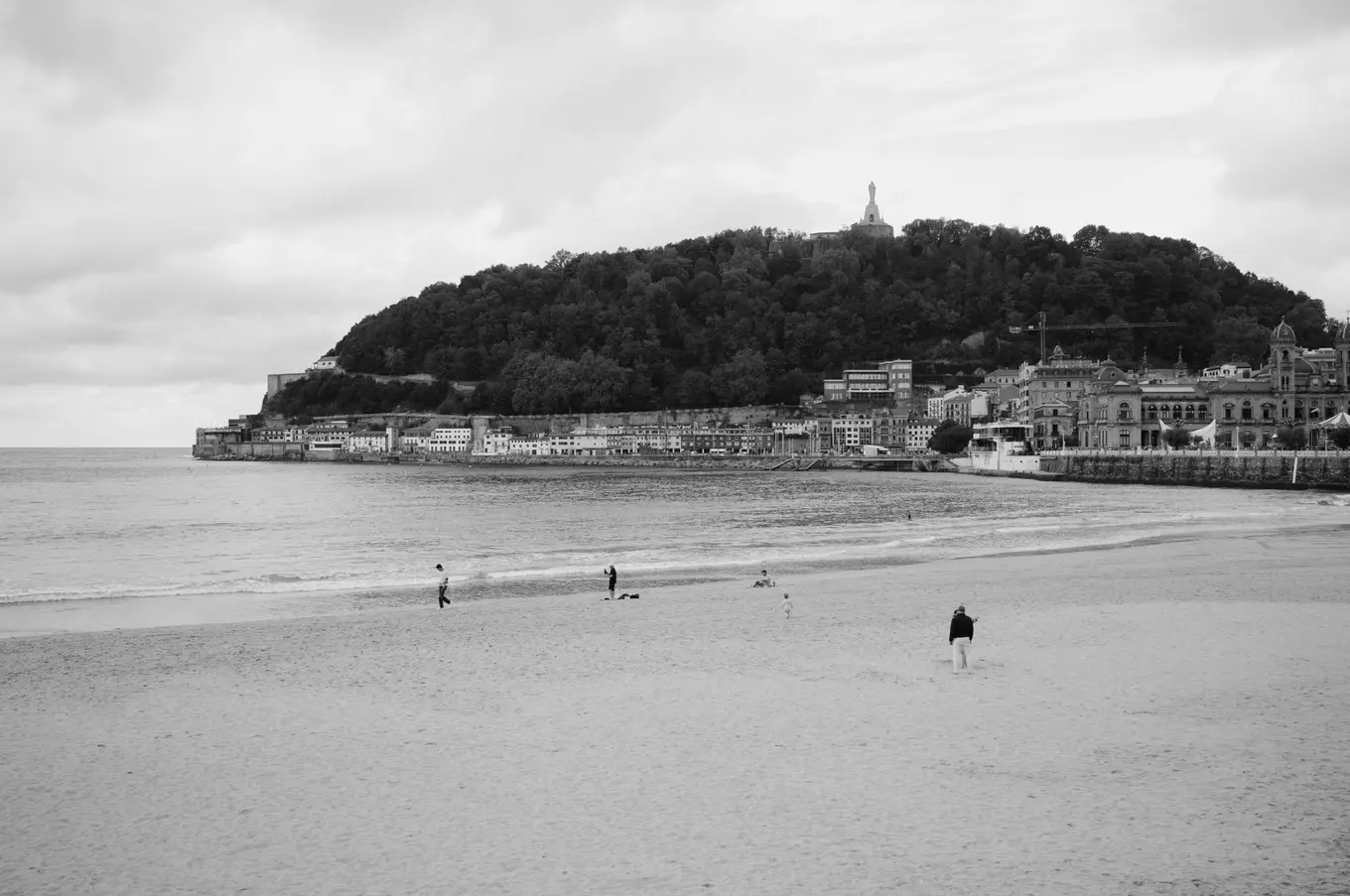 A few figures on a wide, empty beach, a wooded hill with a statue on top rising over the old town across the bay