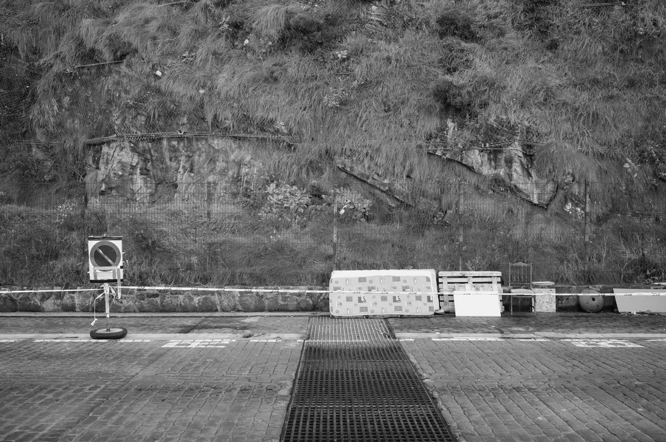 A mattress, a pallet and a chair set out along a low wall below a scrubby hillside, a no-entry sign on a post beside them