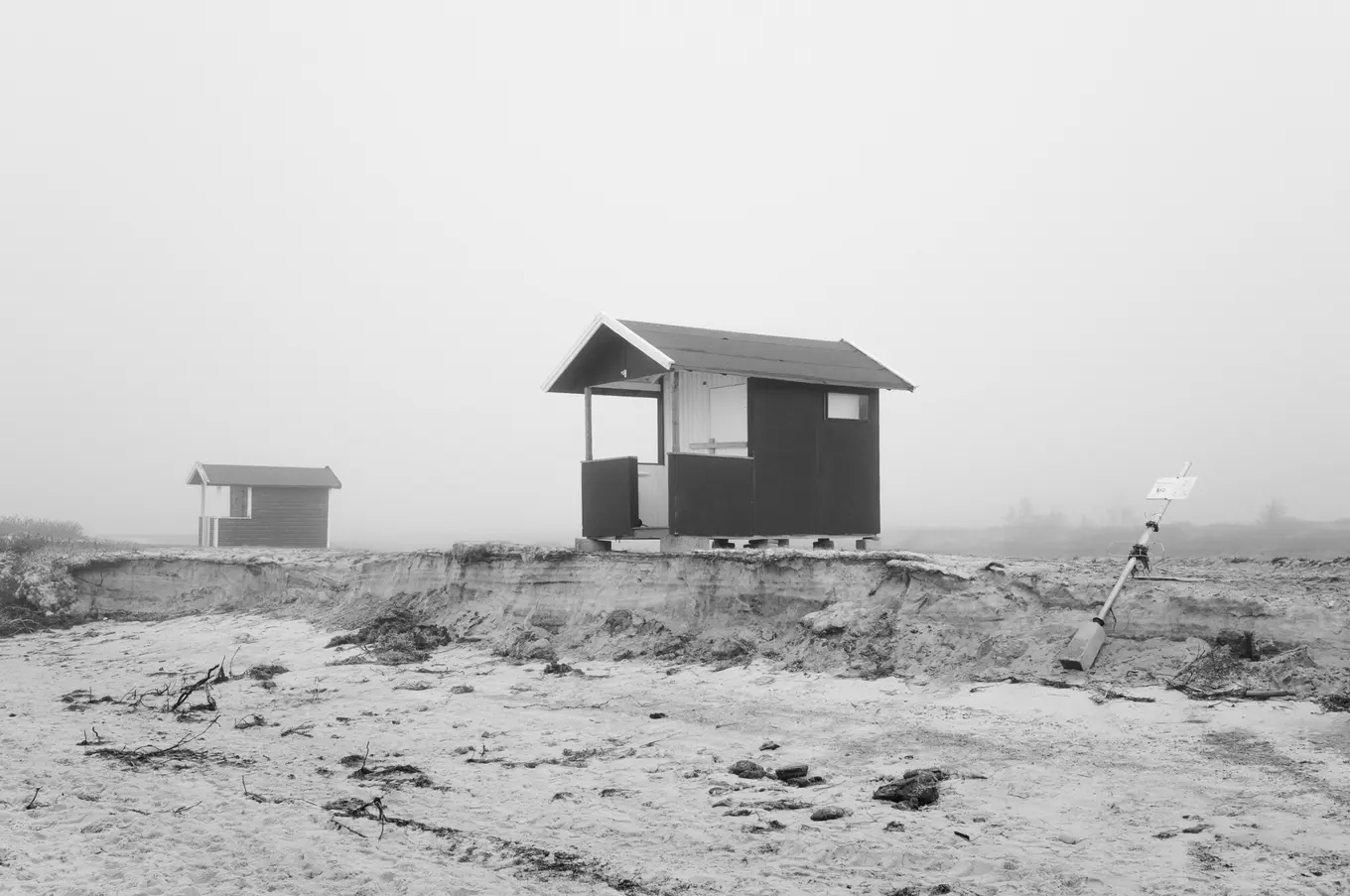 A dark hut and a distant one across flat sand, a sign leaning at the right