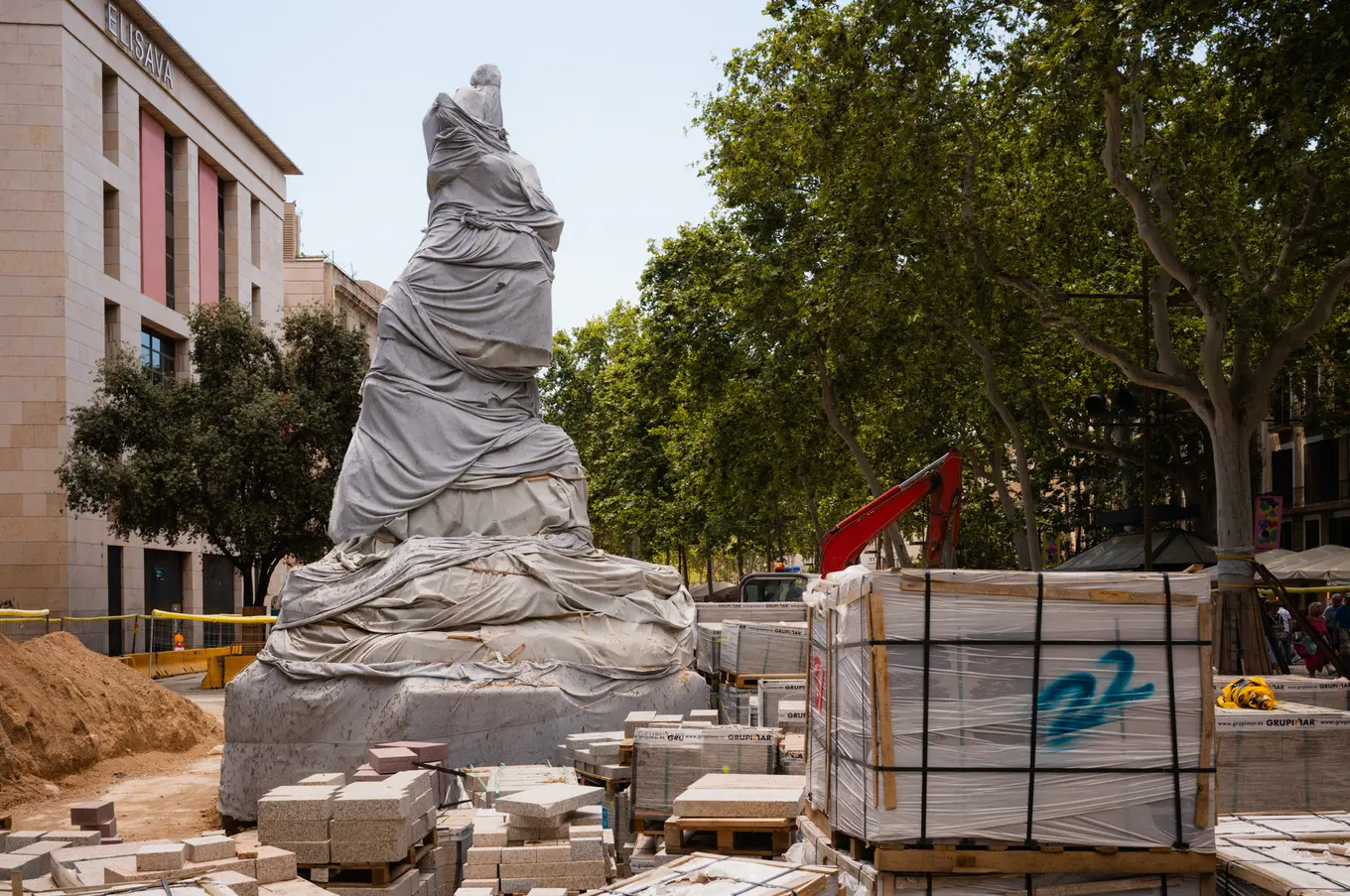 A monument wrapped in grey sheeting, standing over pallets of paving stones on a boulevard under repair