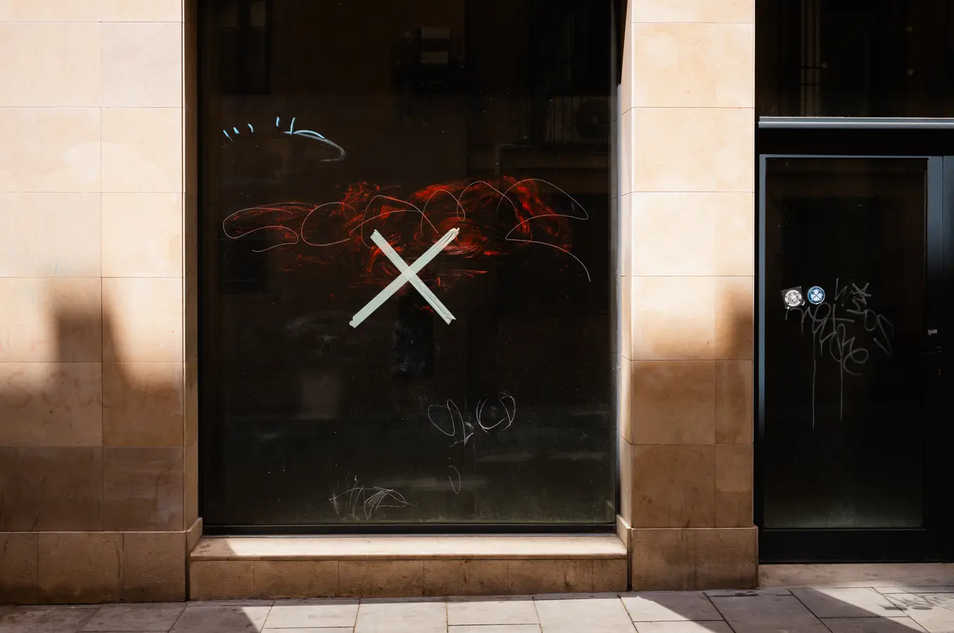 A tall dark shopfront window taped over with a white cross, red graffiti showing faintly behind the glass