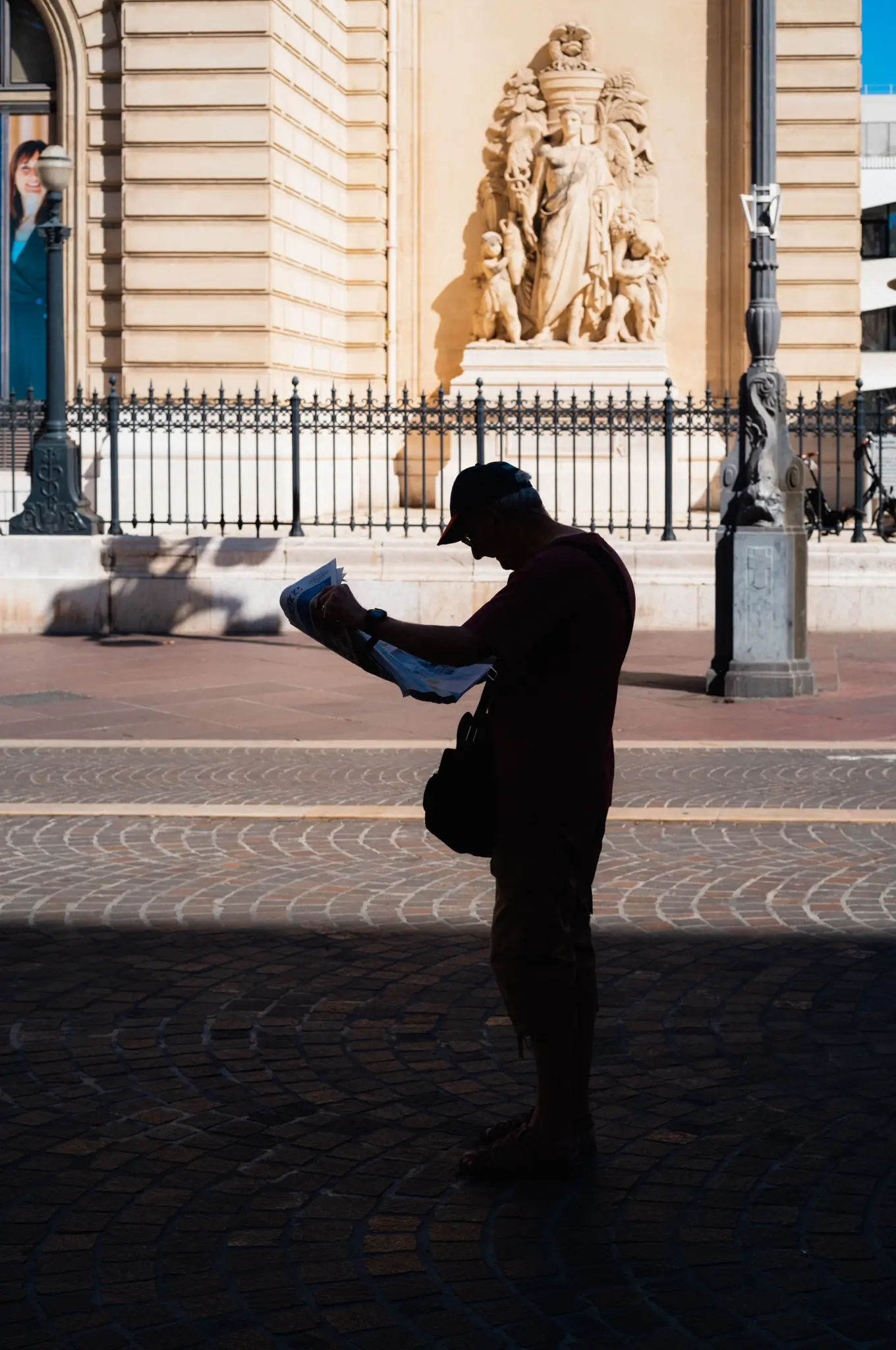 A man in silhouette reading a folded map in deep shadow, a sunlit stone relief behind an iron railing across the street