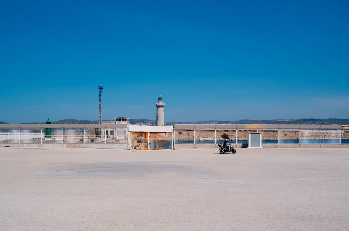 A lone scooter crossing a wide empty quay, a lighthouse and a fenced harbour wall under a clear blue sky