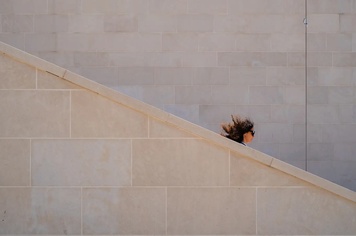 A woman's head and windblown hair just showing above the diagonal edge of a pale stone staircase wall