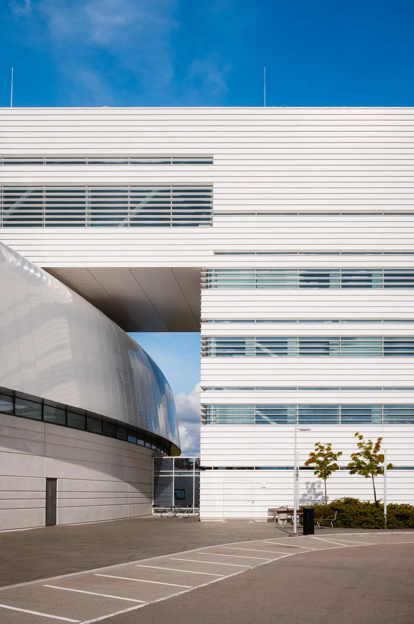 A white slatted office block bridging toward the metal ring hall, an empty parking lot below