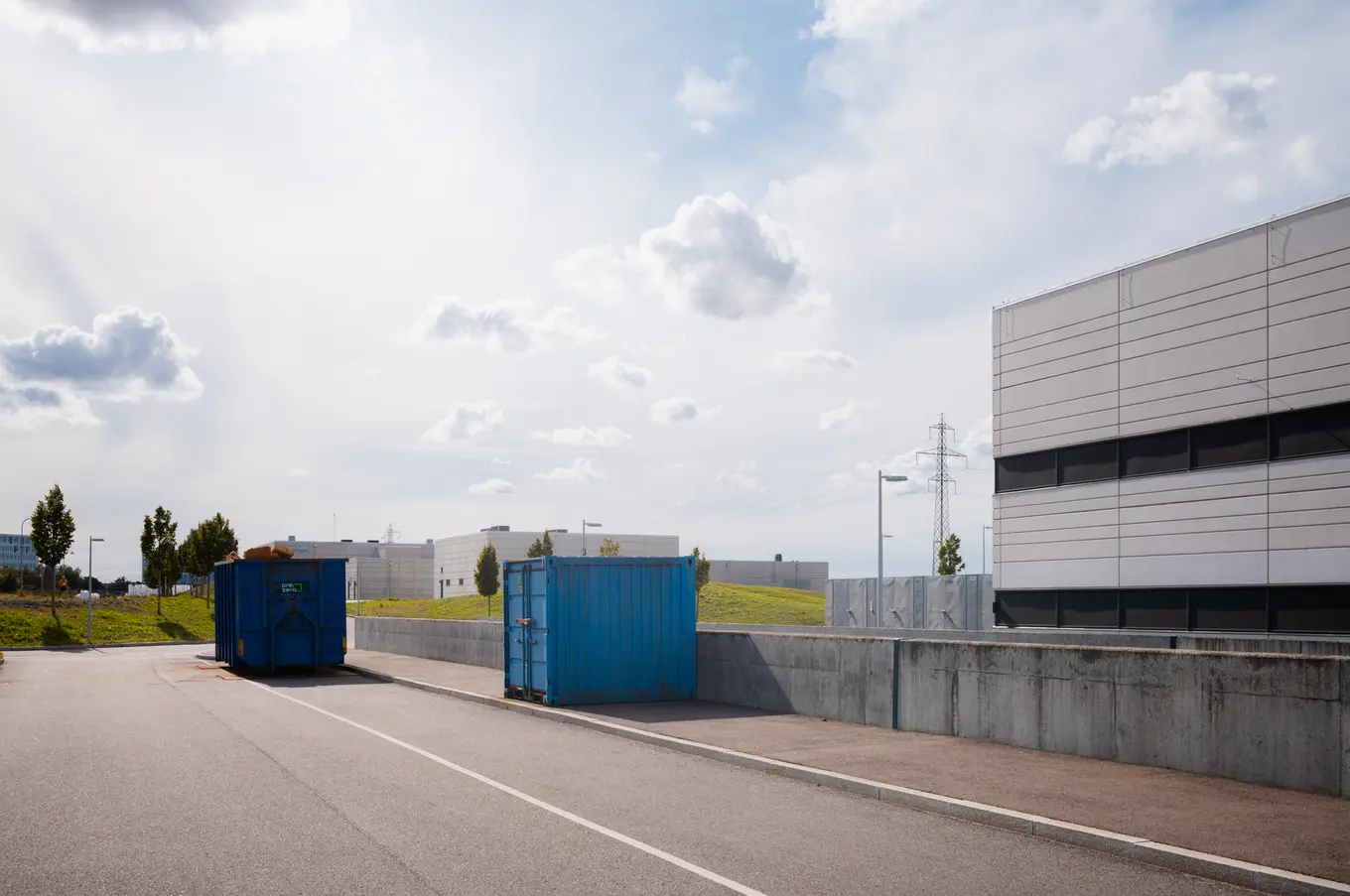 Two blue containers on a service road below a grass bank, a pylon on the horizon