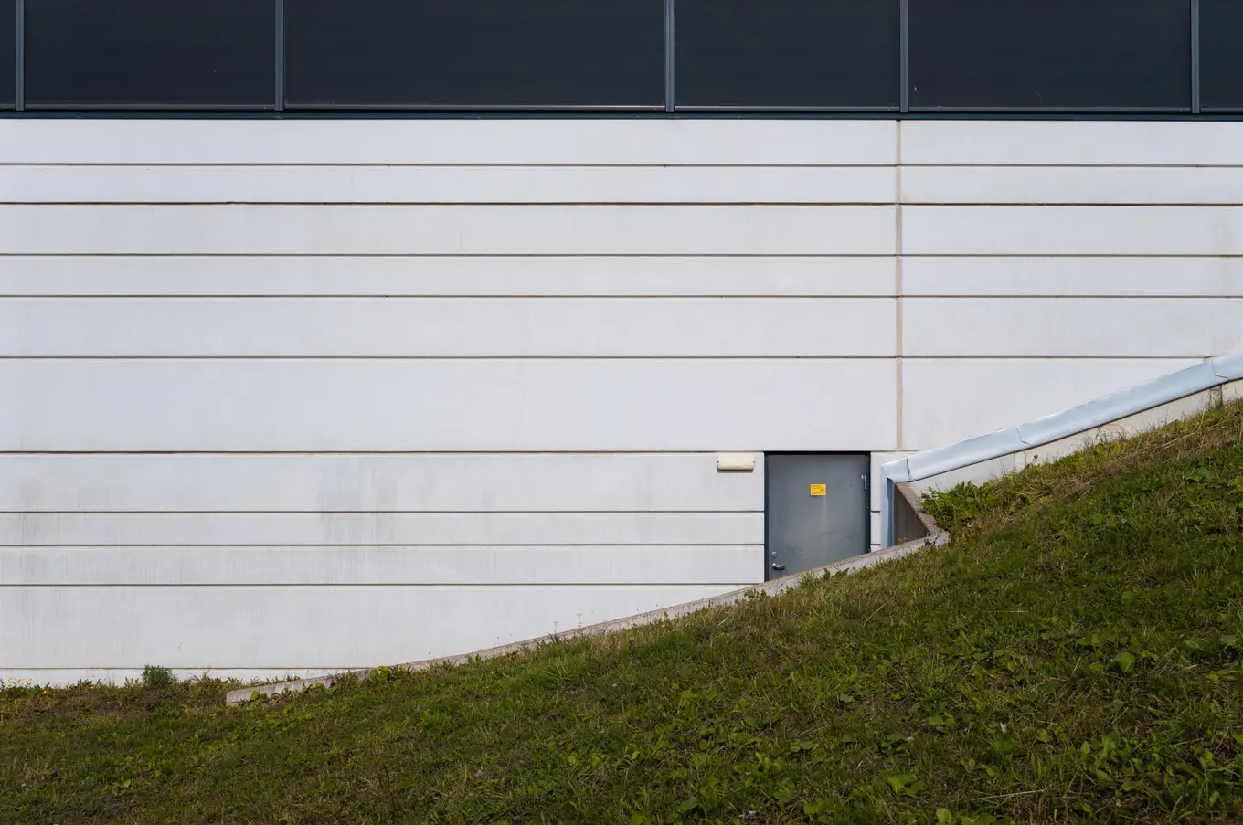 A grey door in a ribbed white wall, a grassed bank rising to meet it