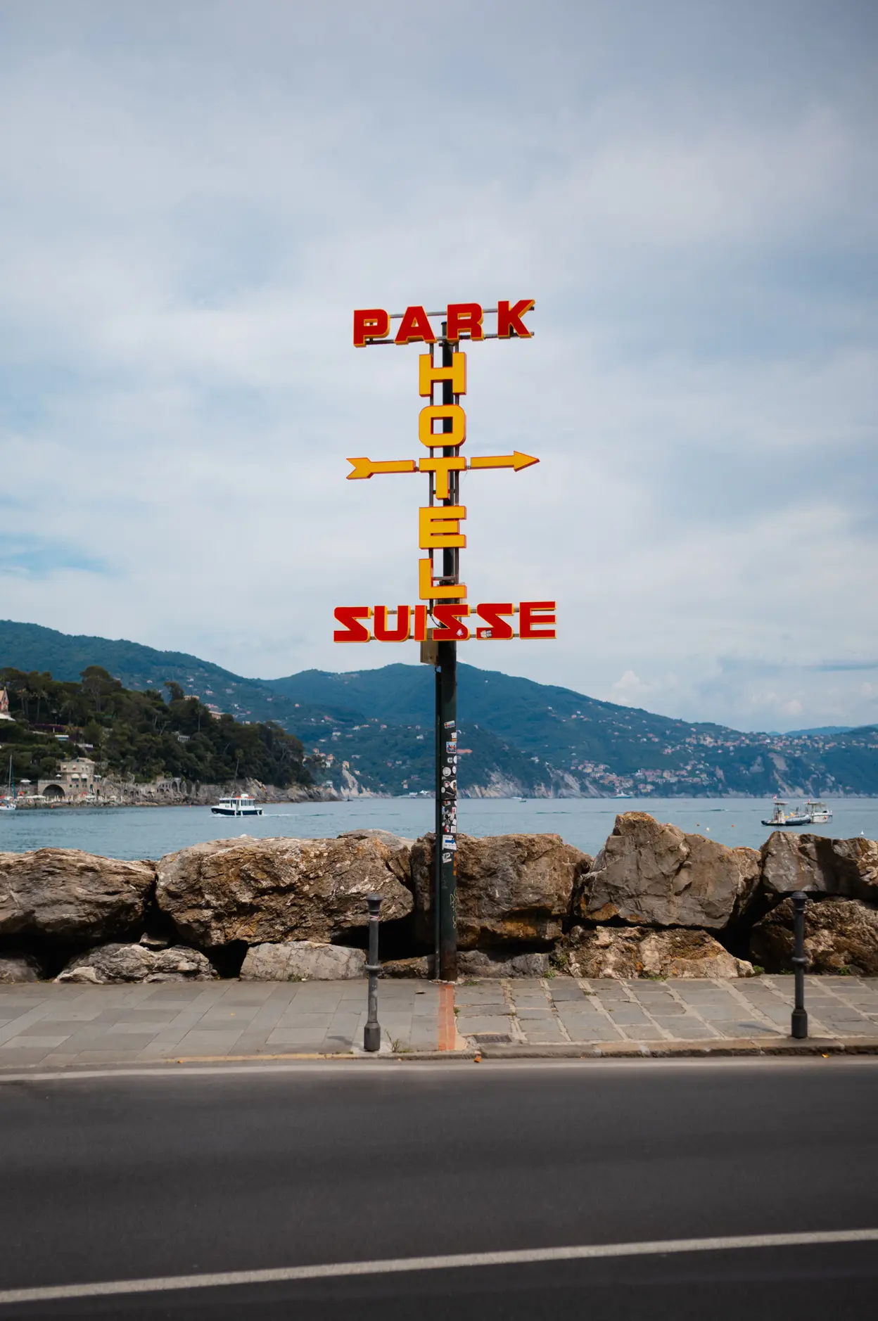 A neon hotel sign on a post above the seafront, hills across the bay
