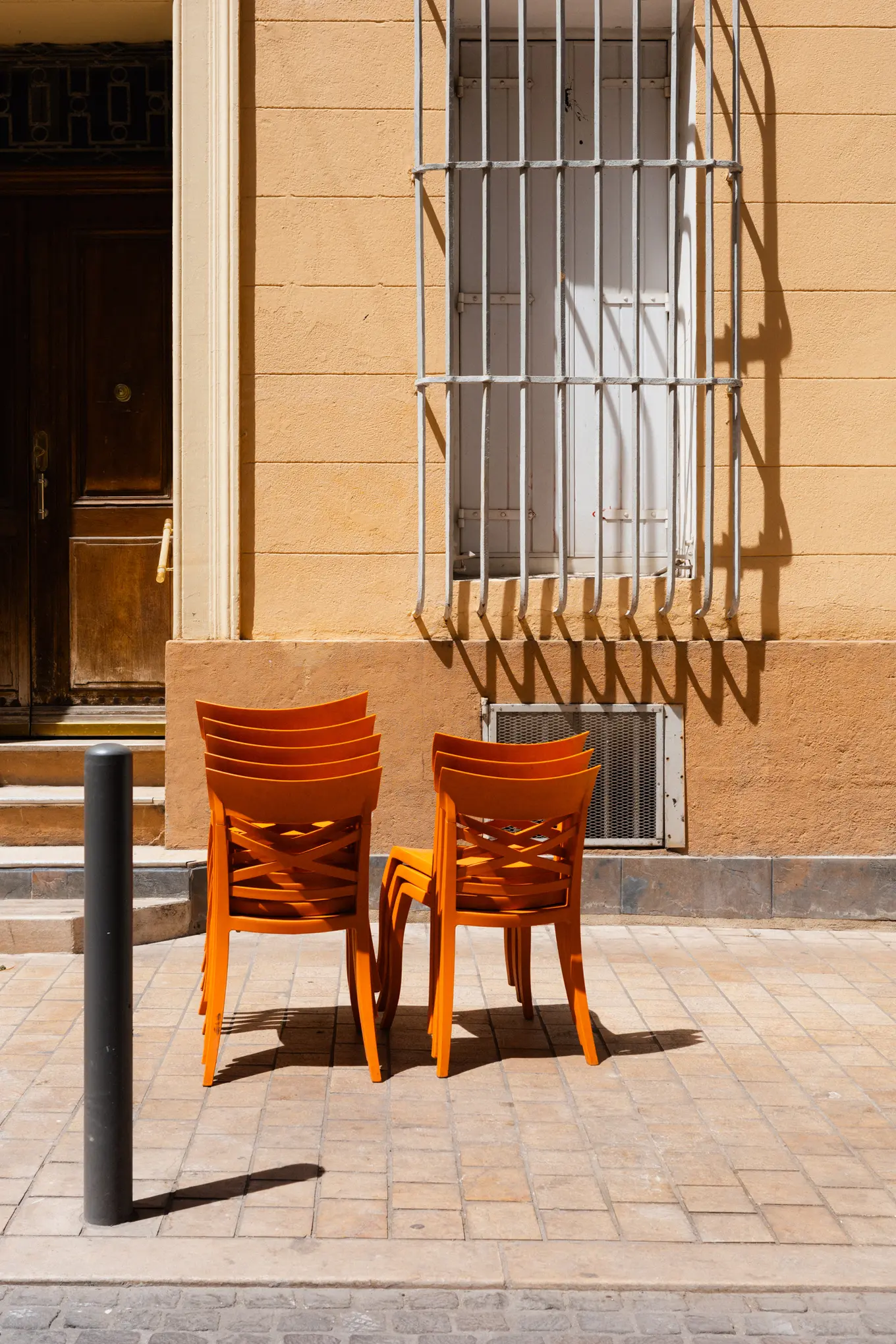 Two stacks of orange chairs on the pavement in front of a yellow house, a barred window casting its shadow down the wall