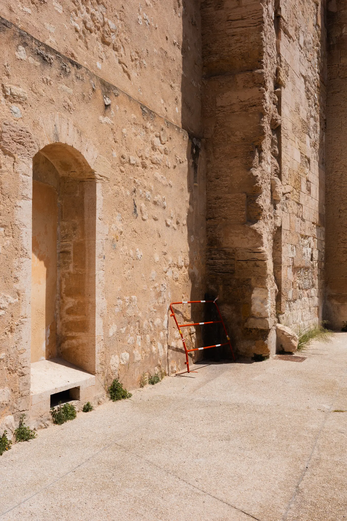 A red and white barrier leaning against the foot of an old sandstone wall beside an empty arched niche